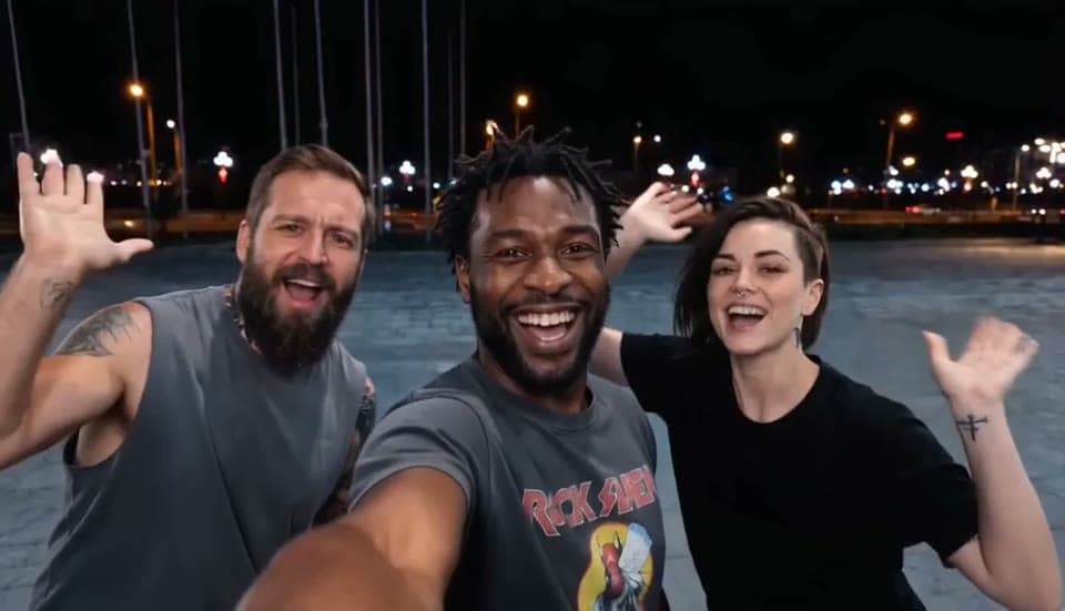 The opening of a Gangnam Style AI sample video at night: a smiling man with short dreadlocks holds the selfie camera in the centre while a bearded man and a dark-haired woman wave on either side